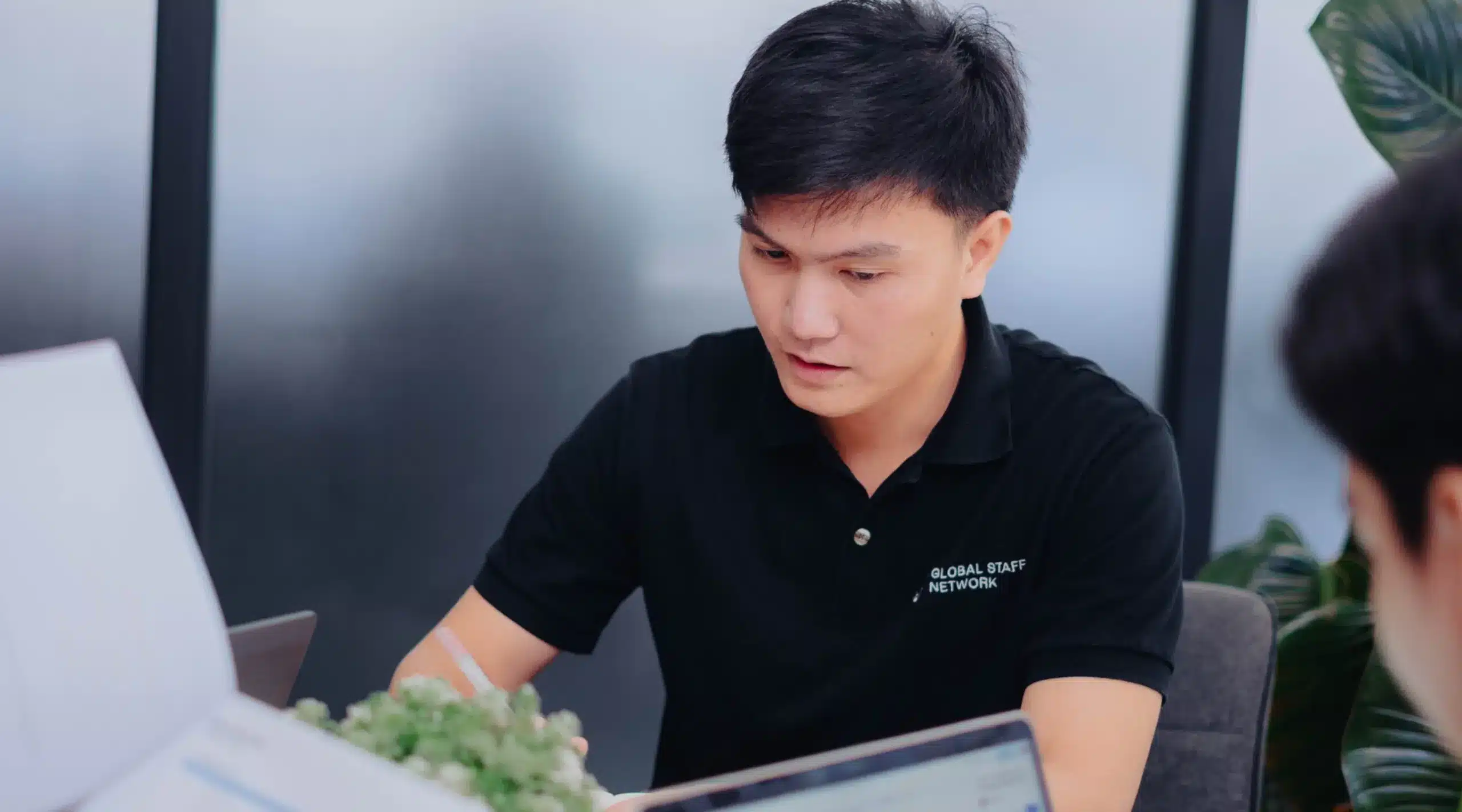 A man wearing a black shirt works at a desk with a laptop, illustrating a global outsourcing network.